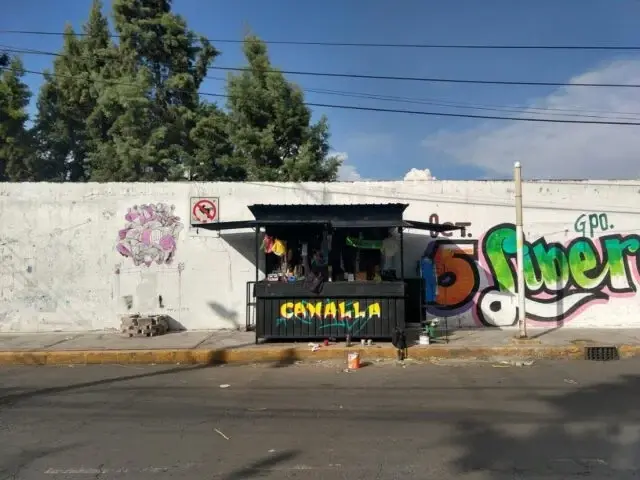 Puesto de calle negro con CANALLA pintado en la fachada, junto a un muro con murales